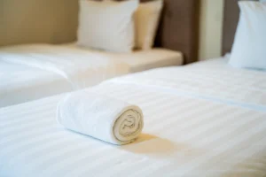 Close-up of neatly rolled white towel placed on a clean, striped hotel bed with soft pillows in the background.