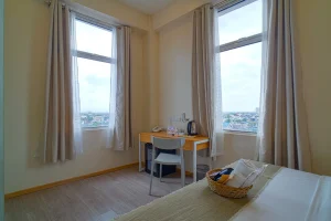 Bright hotel room corner with two large windows, a wooden desk, and a comfortable bed with a towel basket.