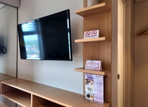 Wall-mounted flat-screen TV with wooden shelf and hotel information cards in a modern guest room.